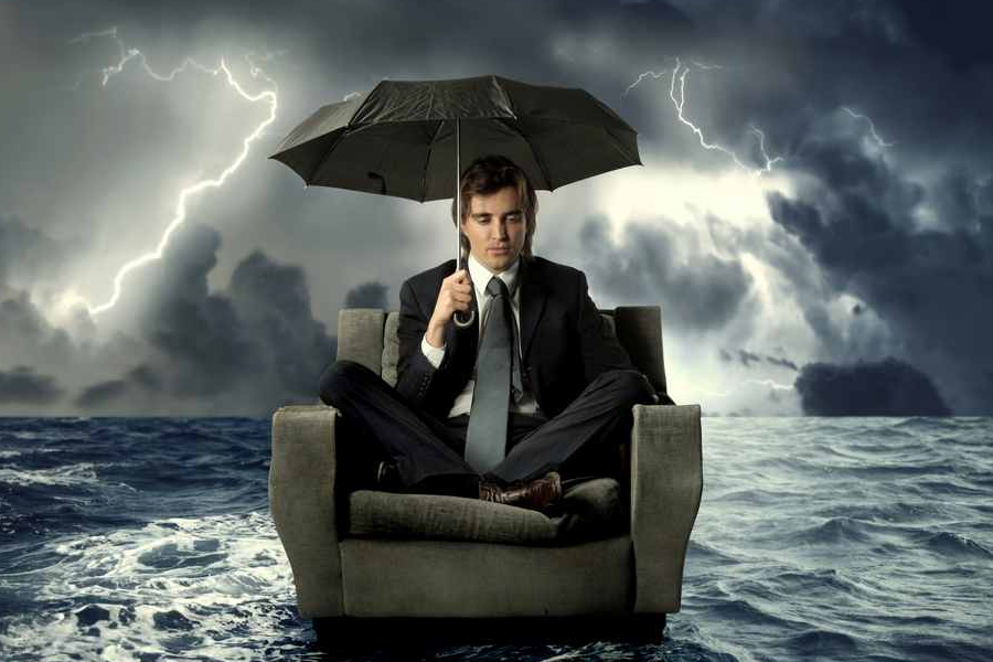 A business man holding an umbrella and sitting on a recliner in the middle of a tumultuous ocean and storm.